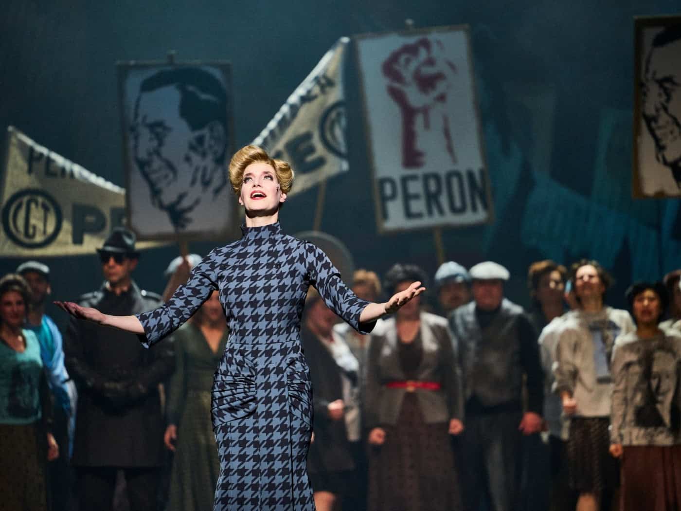 Bühnenfoto aus dem Musical Evita an der Staatsoperette Dresden: Sybille Lambrich steht als Eva Perón in einem blau-schwarz gemusterten Kleid mit ausgebreiteten Armen auf der Bühne, im Hintergrund das Ensemble mit Perón-Protestschildern.