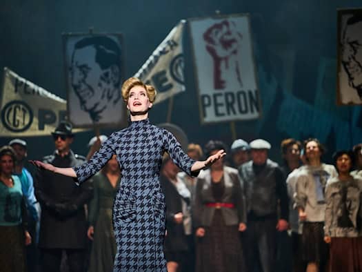 Bühnenfoto aus dem Musical Evita an der Staatsoperette Dresden: Sybille Lambrich steht als Eva Perón in einem blau-schwarz gemusterten Kleid mit ausgebreiteten Armen auf der Bühne, im Hintergrund das Ensemble mit Perón-Protestschildern.