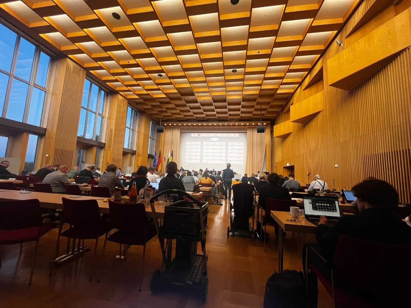 Blick von hinten in den mit Holz verkleideten Plenarsaal des Dresdner Rathauses. Die Stadträte sitzen an ihren Tischen mit Laptops, im Hintergrund ist das Podium mit Präsentationsleinwand sowie den Flaggen von Deutschland und Sachsen zu sehen.