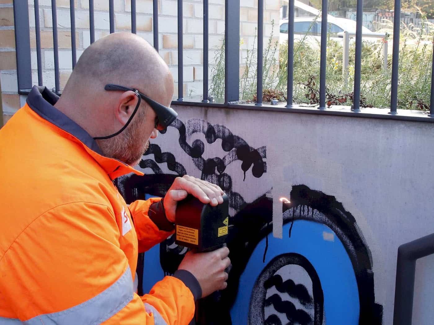 Nahaufnahme eines Mitarbeiters der Stadtreinigung mit Schutzbrille und orangefarbener Warnjacke. Er führt ein Lasergerät dicht an eine Wand, wo ein kleiner Lichtpunkt die schwarze und blaue Sprühfarbe präzise vom Untergrund löst.