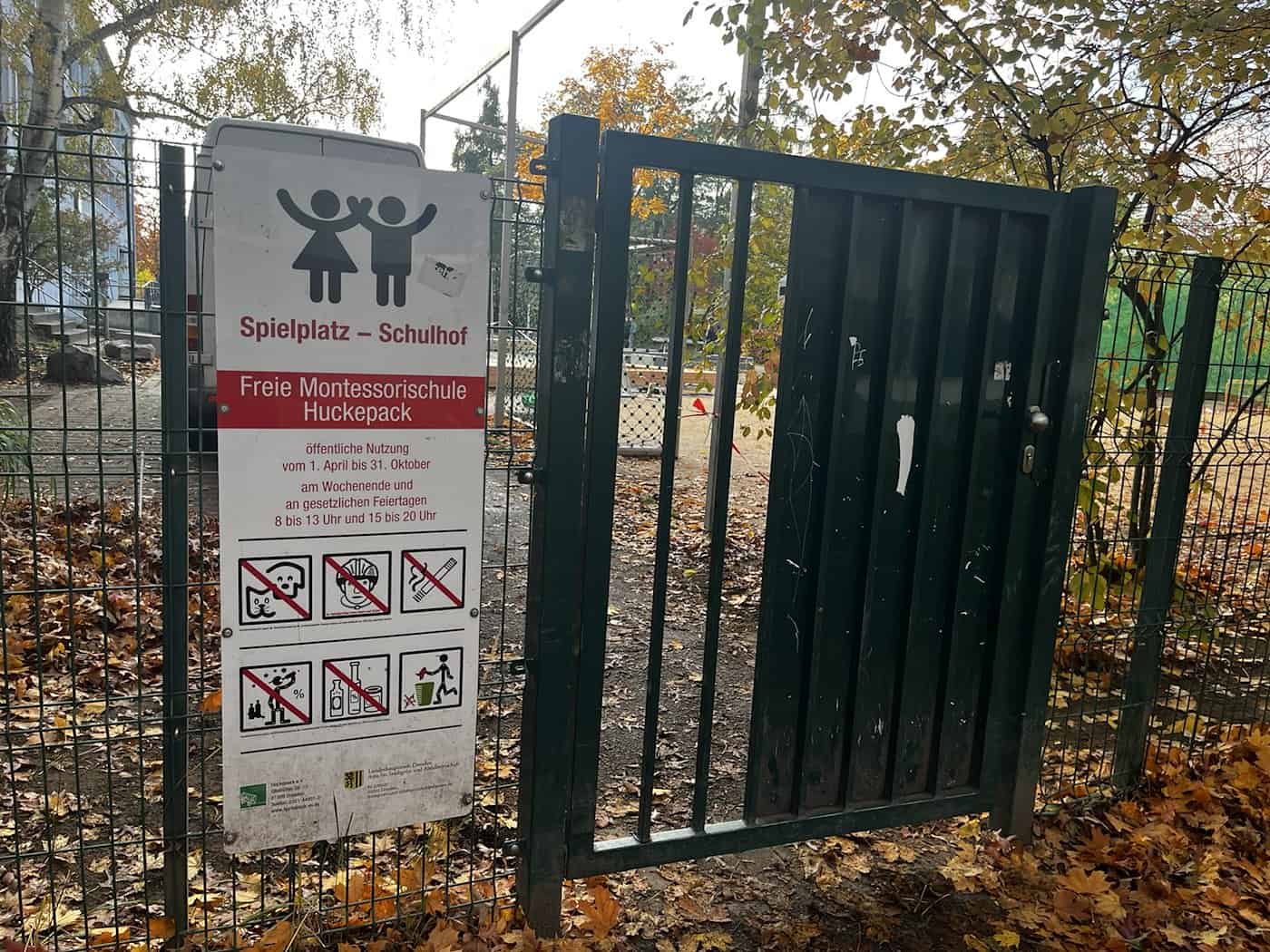 Nahaufnahme des Eingangstors der Freien Montessorischule Huckepack in Dresden. Ein weiß-rotes Schild informiert über die öffentliche Nutzung als Spielplatz an Wochenenden und Feiertagen, umgeben von herbstlich buntem Laub.