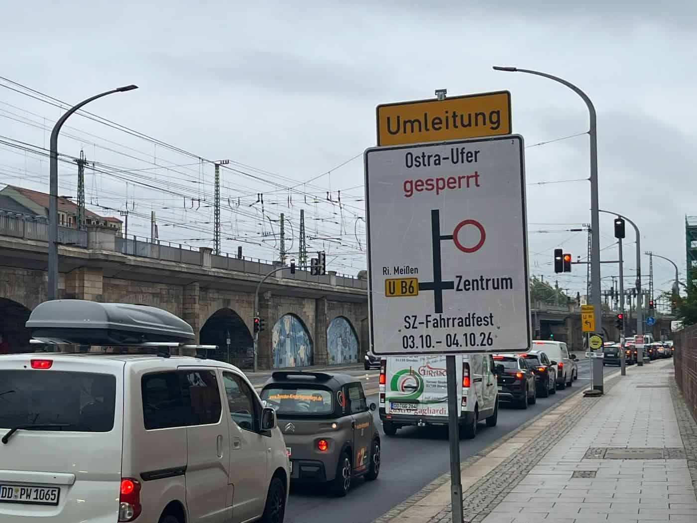 Ein weiß-gelbes Umleitungsschild kündigt die Sperrung des Ostra-Ufers für das SZ-Fahrradfest vom 3. und 4.10.26 an. Daneben stauen sich Autos vor einer Fußgängerampel.