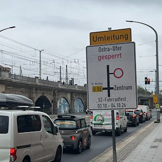 Ein weiß-gelbes Umleitungsschild kündigt die Sperrung des Ostra-Ufers für das SZ-Fahrradfest vom 3. und 4.10.26 an. Daneben stauen sich Autos vor einer Fußgängerampel.