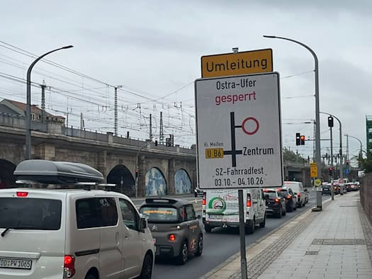 Ein weiß-gelbes Umleitungsschild kündigt die Sperrung des Ostra-Ufers für das SZ-Fahrradfest vom 3. und 4.10.26 an. Daneben stauen sich Autos vor einer Fußgängerampel.