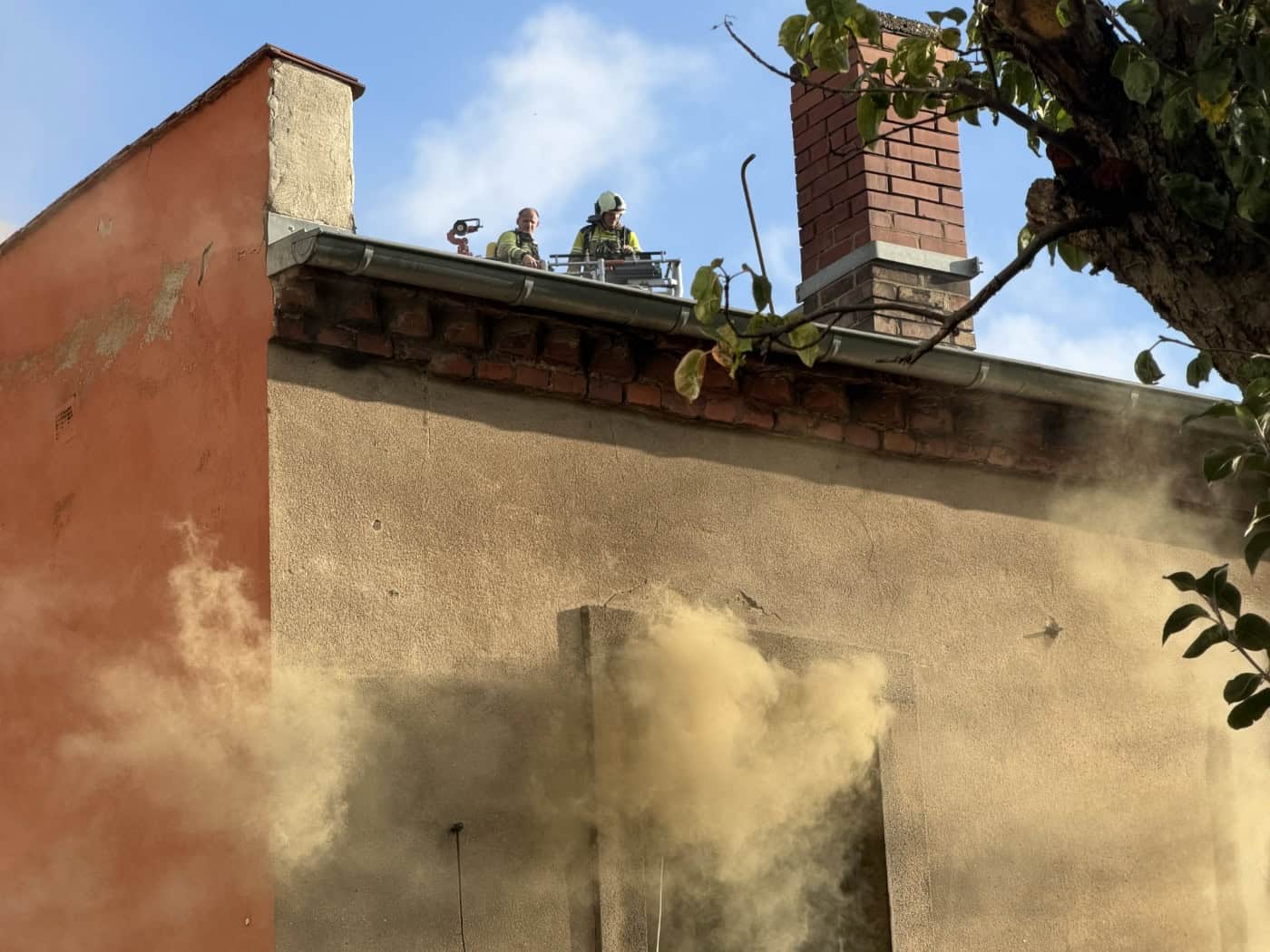 Symbolbild: Dichter, gelblicher Rauch quillt aus einem geöffneten Obergeschossfenster eines Wohnhauses. Im Hintergrund sind Einsatzkräfte der Feuerwehr auf einer Drehleiter am Dachbereich zu erkennen, im Vordergrund ragen Baumäste ins Bild.