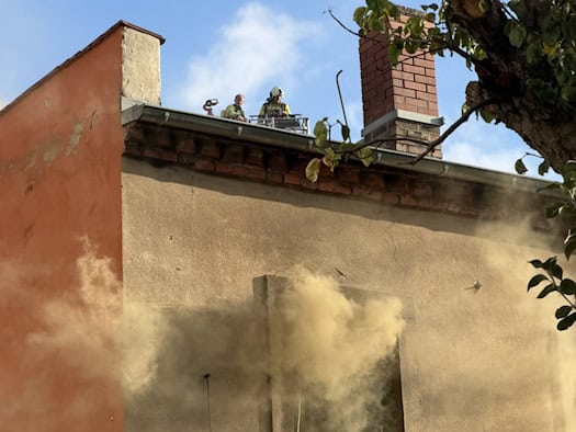Symbolbild: Dichter, gelblicher Rauch quillt aus einem geöffneten Obergeschossfenster eines Wohnhauses. Im Hintergrund sind Einsatzkräfte der Feuerwehr auf einer Drehleiter am Dachbereich zu erkennen, im Vordergrund ragen Baumäste ins Bild.