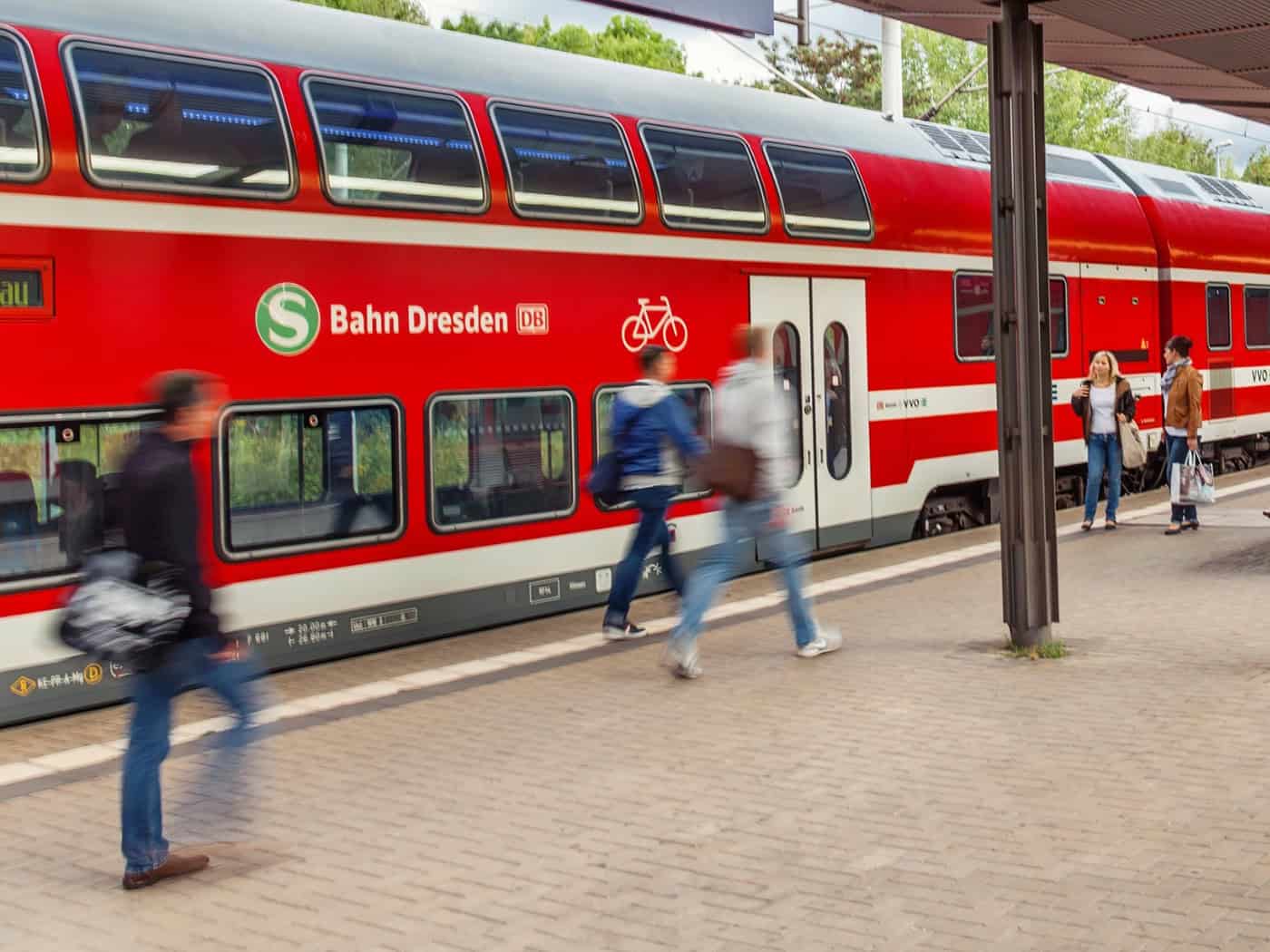 Rote Doppelstock-Züge der S-Bahn Dresden stehen an einem überdachten Bahnsteig. Passagiere gehen über den Bahnsteig und steigen in die Bahn ein.