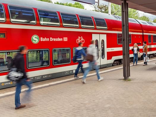 Rote Doppelstock-Züge der S-Bahn Dresden stehen an einem überdachten Bahnsteig. Passagiere gehen über den Bahnsteig und steigen in die Bahn ein.