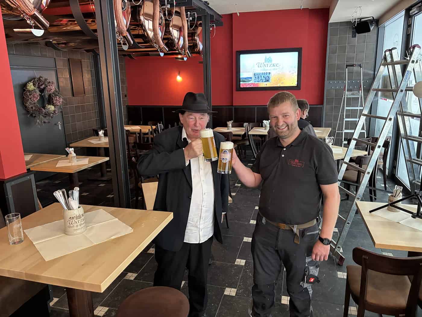 Braumeister Jens Berndt (rechts) und Künstler Holger John mit Hut und dunklem Sakko (links) stehen im modernisierten Gastraum des Watzke am Goldenen Reiter und stoßen mit zwei gefüllten Biergläsern an. Im Hintergrund stehen Tische und eine Anlegeleiter.