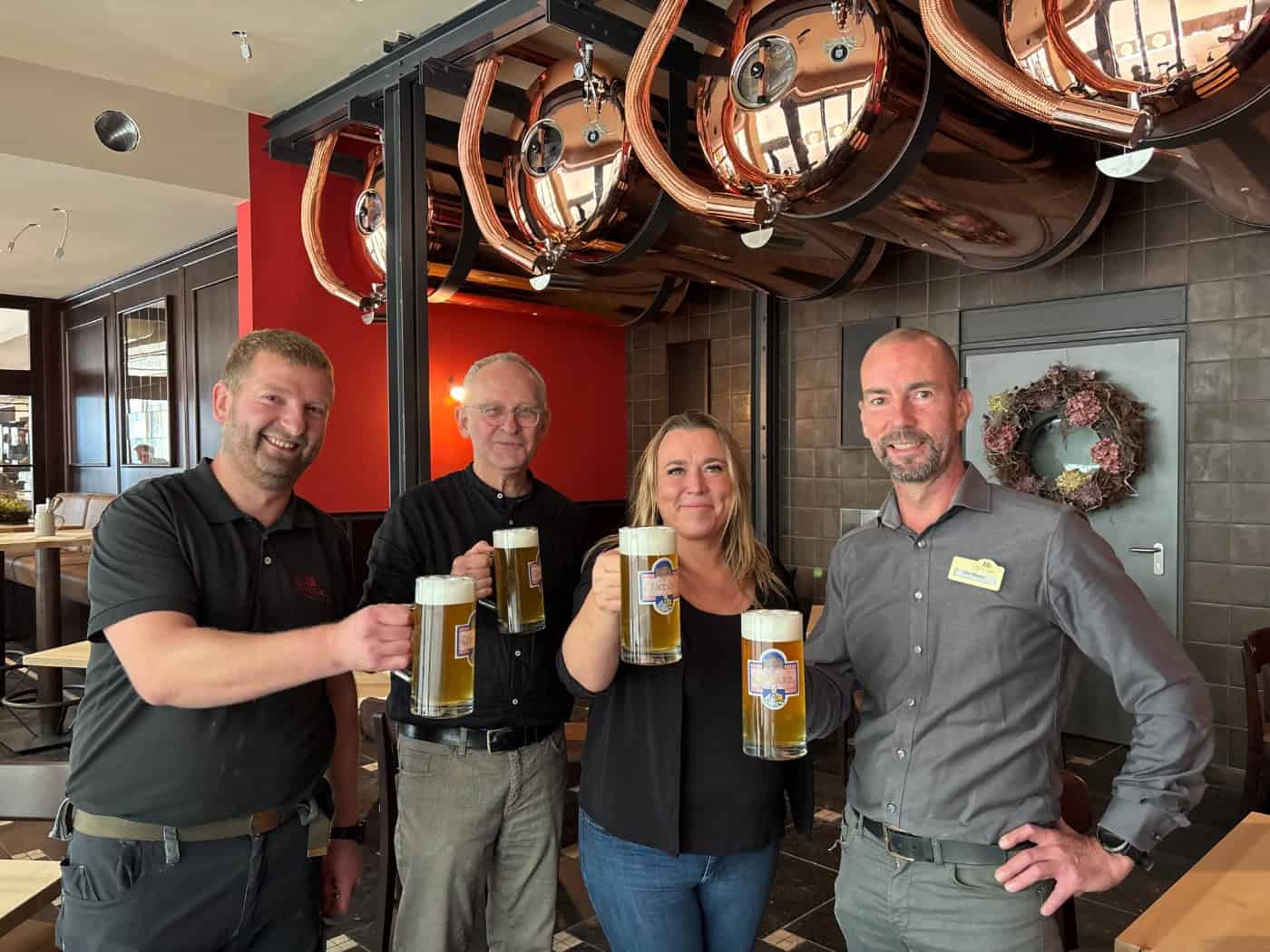Vier Personen stehen nebeneinander im Gastraum und stoßen mit voll gefüllten Bierkrügen an: v.l.n.r. Braumeister Jens Berndt, Architekt Holger Stetzler, Geschäftsführerin Nicole Rothländer und Restaurantleiter Mike Böhme unter hängenden Kupfertanks.