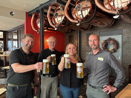 Vier Personen stehen nebeneinander im Gastraum und stoßen mit voll gefüllten Bierkrügen an: v.l.n.r. Braumeister Jens Berndt, Architekt Holger Stetzler, Geschäftsführerin Nicole Rothländer und Restaurantleiter Mike Böhme unter hängenden Kupfertanks.