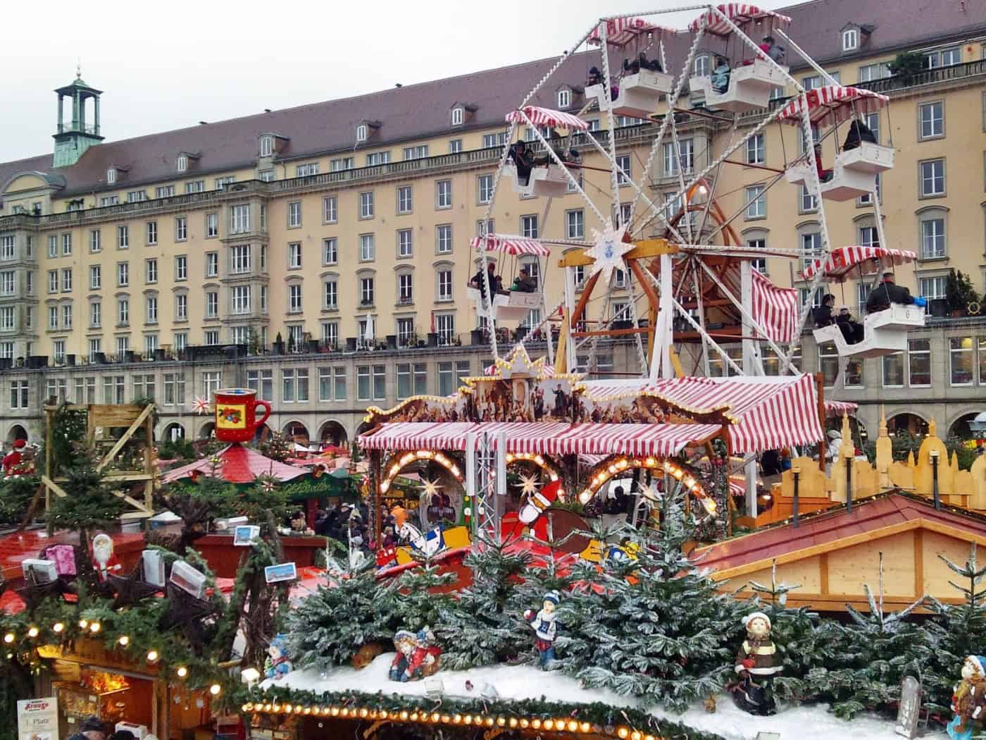 Festlich geschmückte Verkaufsstände auf dem Dresdner Striezelmarkt mit einem historischen Riesenrad, Lichterketten und Tannengrün vor einer großen Häuserfassade.
