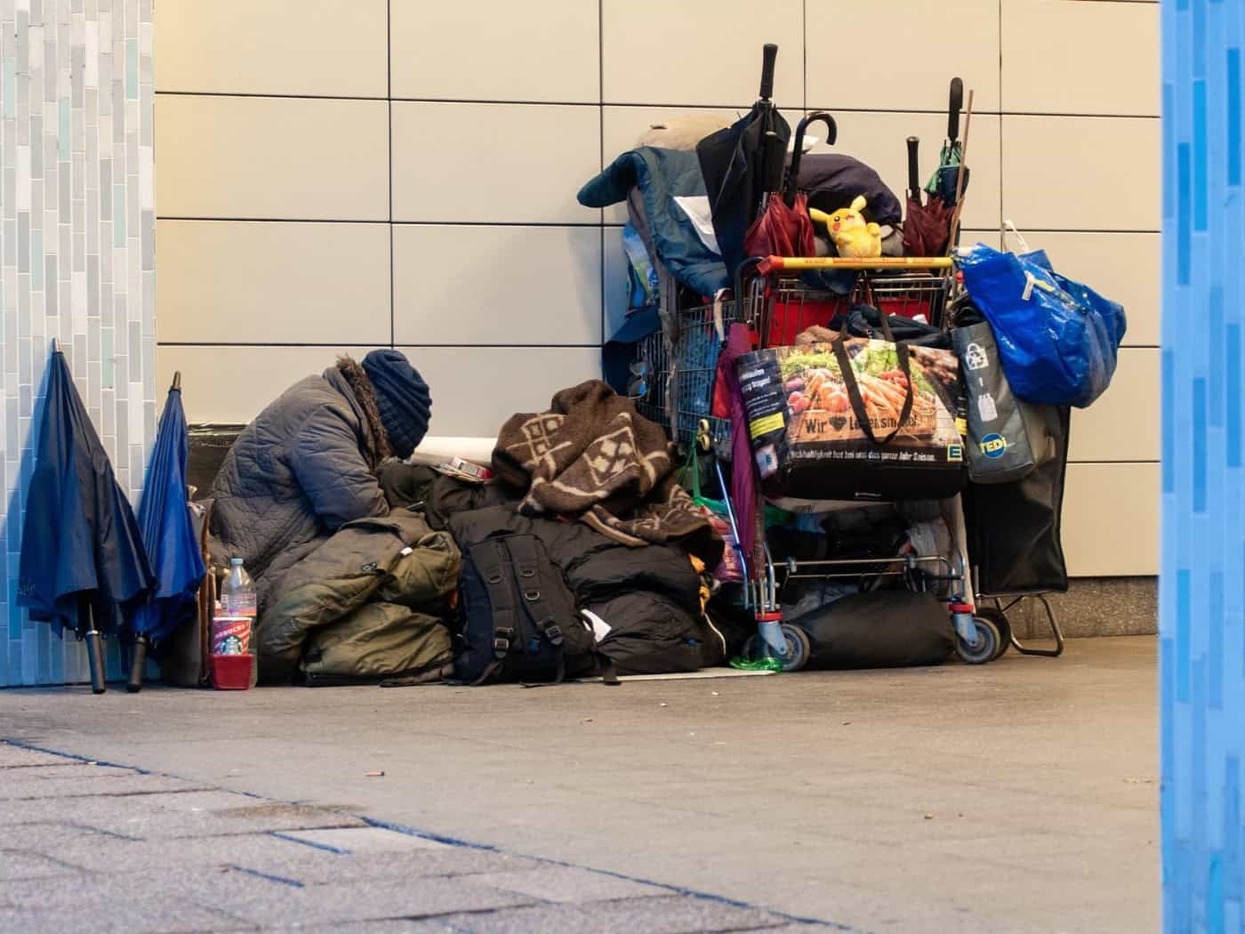 Symbolbild zu Wohnungslosigkeit: Eine Person sitzt eingekuschelt in dicke Decken und warme Kleidung auf dem Boden vor einer Hauswand. Neben ihr steht ein vollgepackter Einkaufswagen mit Taschen, Regenschirmen und Decken.