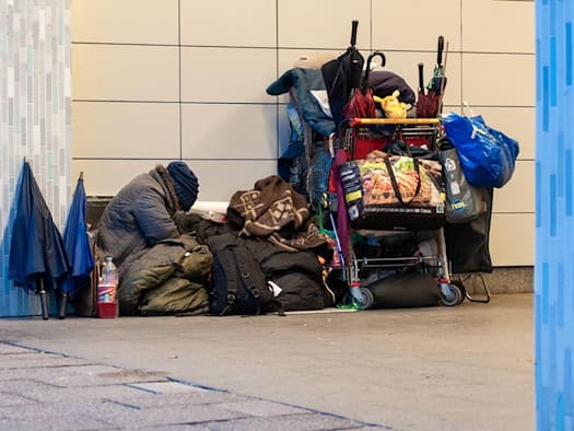 Symbolbild zu Wohnungslosigkeit: Eine Person sitzt eingekuschelt in dicke Decken und warme Kleidung auf dem Boden vor einer Hauswand. Neben ihr steht ein vollgepackter Einkaufswagen mit Taschen, Regenschirmen und Decken.