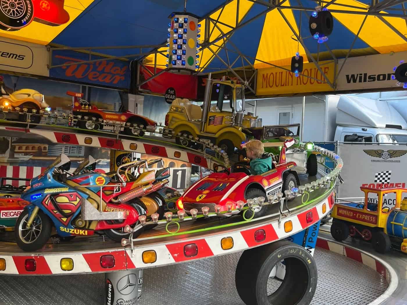 Ein blondes Kind fährt in einem roten Spielzeug-Rennauto auf einer angehobenen Kurvenbahn eines bunten Kinderkarussells unter einem gelb-blauen Zeltdach. Im Vordergrund steht ein Minimotorrad mit Superman-Aufschrift.