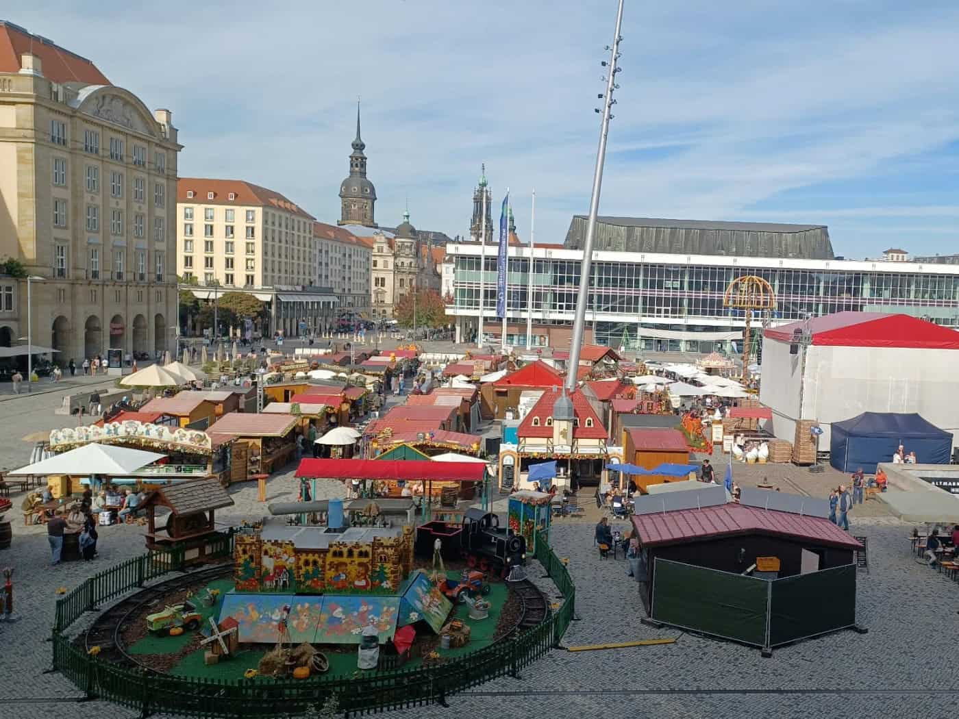 Blick von oben auf den Dresdner Altmarkt während des Herbstmarktes: Zahlreiche rot gedeckte Marktbuden, eine kleine Kindereisenbahn im eingezäunten Vordergrund und der Kulturpalast im Hintergrund unter sonnigem Himmel.