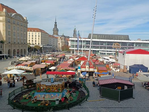 Blick von oben auf den Dresdner Altmarkt während des Herbstmarktes: Zahlreiche rot gedeckte Marktbuden, eine kleine Kindereisenbahn im eingezäunten Vordergrund und der Kulturpalast im Hintergrund unter sonnigem Himmel.