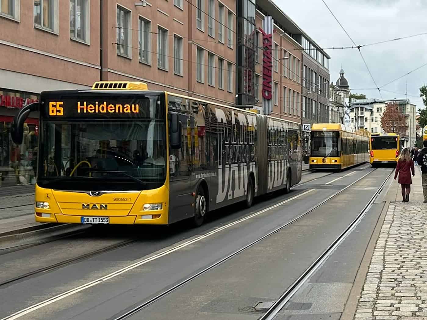Ein gelber DVB-Gelenkbus der Linie 65 mit der Zielanzeige „Heidenau“ fährt auf Schienen durch eine Einkaufsstraße in Dresden. Rechts gehen Passanten auf einem Gehweg, im Hintergrund sind Geschäftshäuser und weitere Fahrzeuge zu sehen.