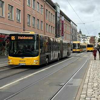 Ein gelber DVB-Gelenkbus der Linie 65 mit der Zielanzeige „Heidenau“ fährt auf Schienen durch eine Einkaufsstraße in Dresden. Rechts gehen Passanten auf einem Gehweg, im Hintergrund sind Geschäftshäuser und weitere Fahrzeuge zu sehen.