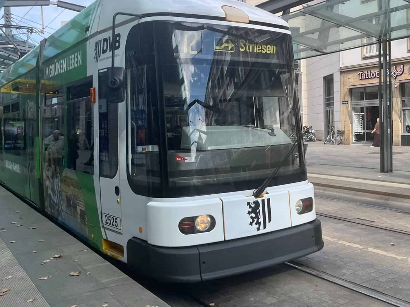 Eine weiß-grüne DVB-Straßenbahn der Linie 12 mit Zielanzeige „Striesen“ steht unter dem Glasdach einer Haltestelle in der Dresdner Innenstadt. Auf dem Bahnsteig im Hintergrund stehen Passanten vor Ladengeschäften.