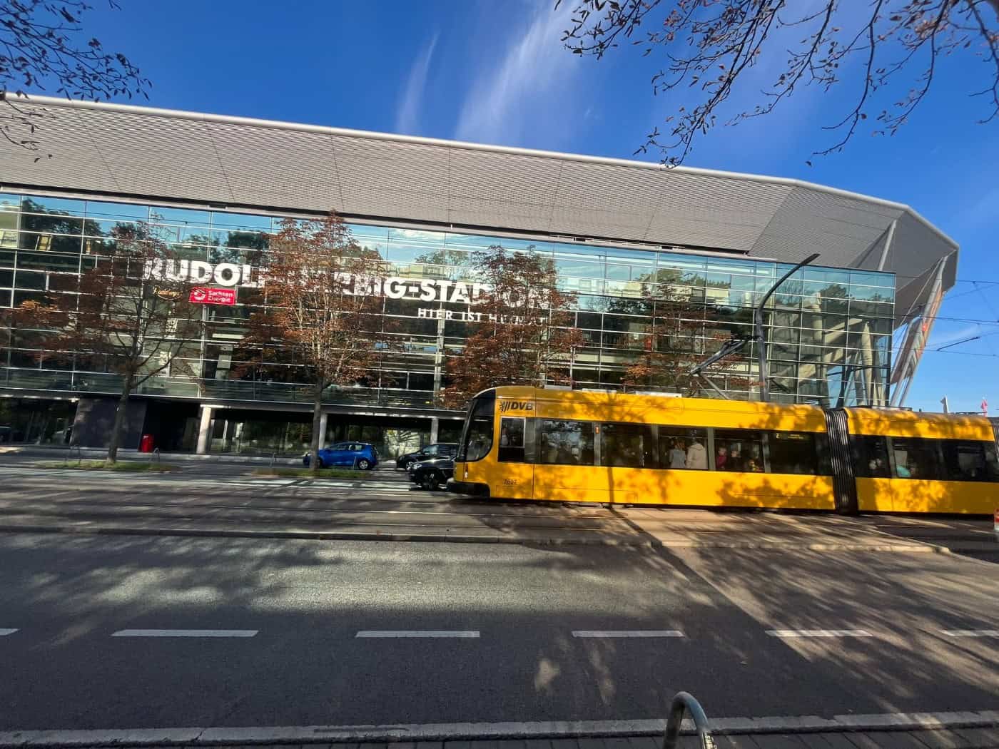 Eine gelbe Straßenbahn der DVB fährt vor der Glasfassade des Rudolf-Harbig-Stadions in Dresden entlang; an der Straße stehen herbstliche Bäume unter einem blauen Himmel mit leichten Wolken.