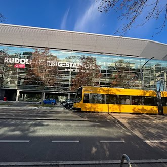 Eine gelbe Straßenbahn der DVB fährt vor der Glasfassade des Rudolf-Harbig-Stadions in Dresden entlang; an der Straße stehen herbstliche Bäume unter einem blauen Himmel mit leichten Wolken.