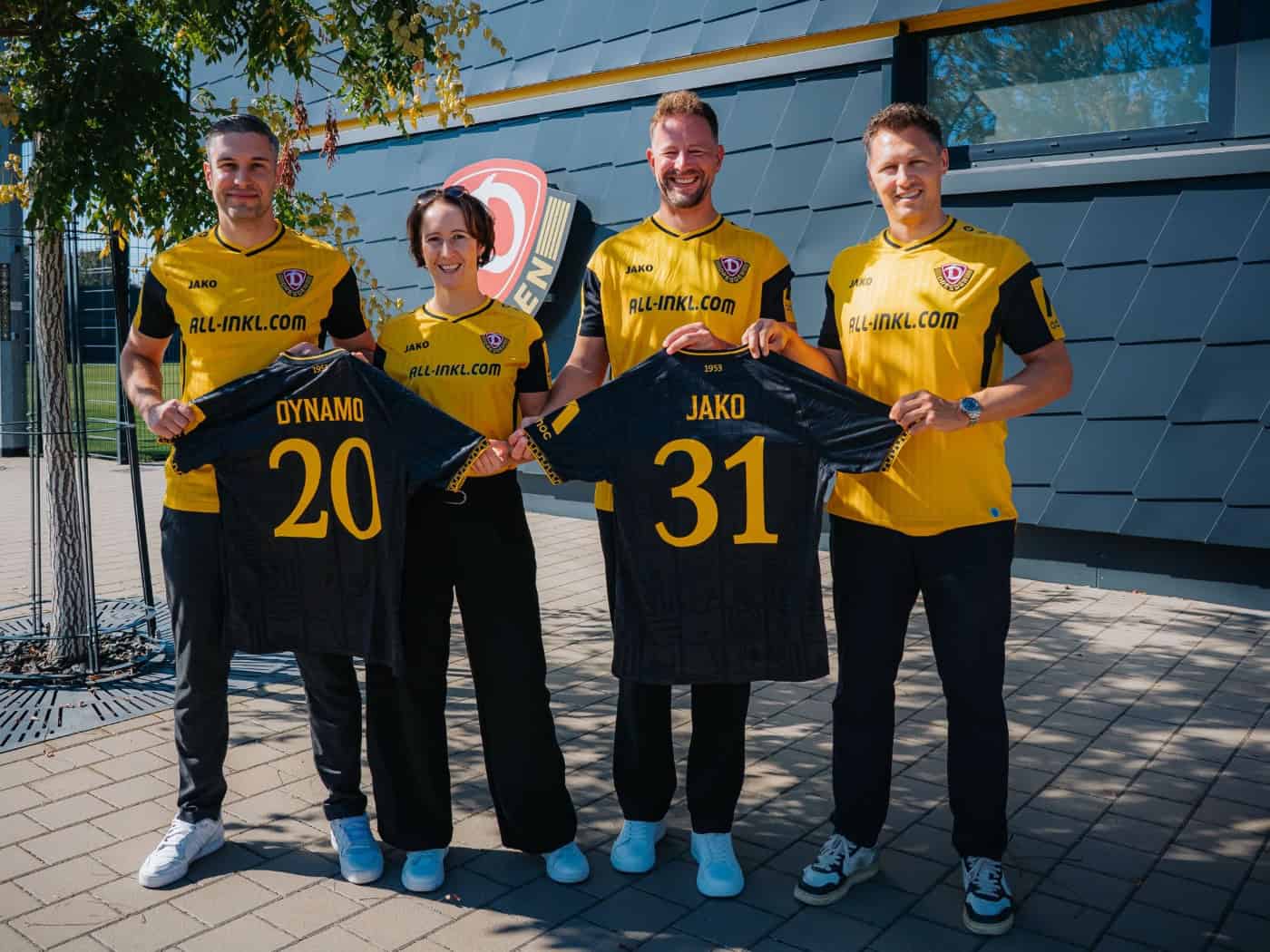Vier Personen in gelb-schwarzen Dynamo-Dresden-Trikots stehen nebeneinander im Freien vor einem Vereinsgebäude. In der Mitte halten sie zwei schwarze Trikots mit der gelben Beflockung „DYNAMO 20“ und „JAKO 31“ für die Vertragsverlängerung in die Kamera.