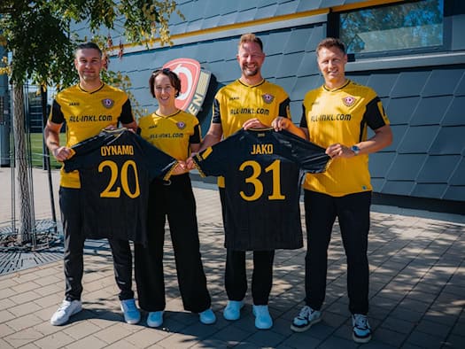 Vier Personen in gelb-schwarzen Dynamo-Dresden-Trikots stehen nebeneinander im Freien vor einem Vereinsgebäude. In der Mitte halten sie zwei schwarze Trikots mit der gelben Beflockung „DYNAMO 20“ und „JAKO 31“ für die Vertragsverlängerung in die Kamera.