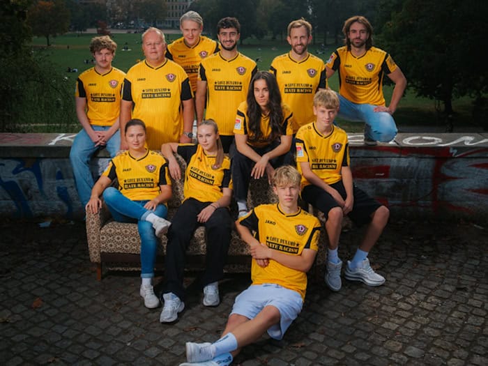 Gruppe von zehn Personen in gelb-schwarzen Dynamo-Dresden-Sondertrikots mit dem Aufdruck „LOVE DYNAMO - HATE RACISM“ posiert im Freien auf einer Steinmauer und einem Sofa. Mit dabei sind Profis, Nachwuchs-, Frauen-Spielerinnen, Vereinsmitarbeiter sowie die Band 01099.