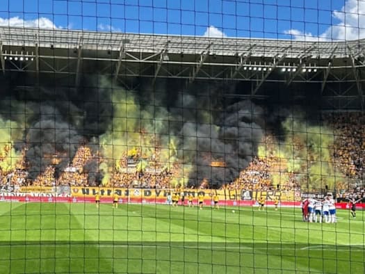 Blick durch ein Fangnetz auf das Spielfeld und die Tribüne im Rudolf-Harbig-Stadion, aus der dichter gelber und schwarzer Rauch von gezündeter Pyrotechnik emporsteigt, während sich die Fußballmannschaften auf dem Rasen aufstellen.