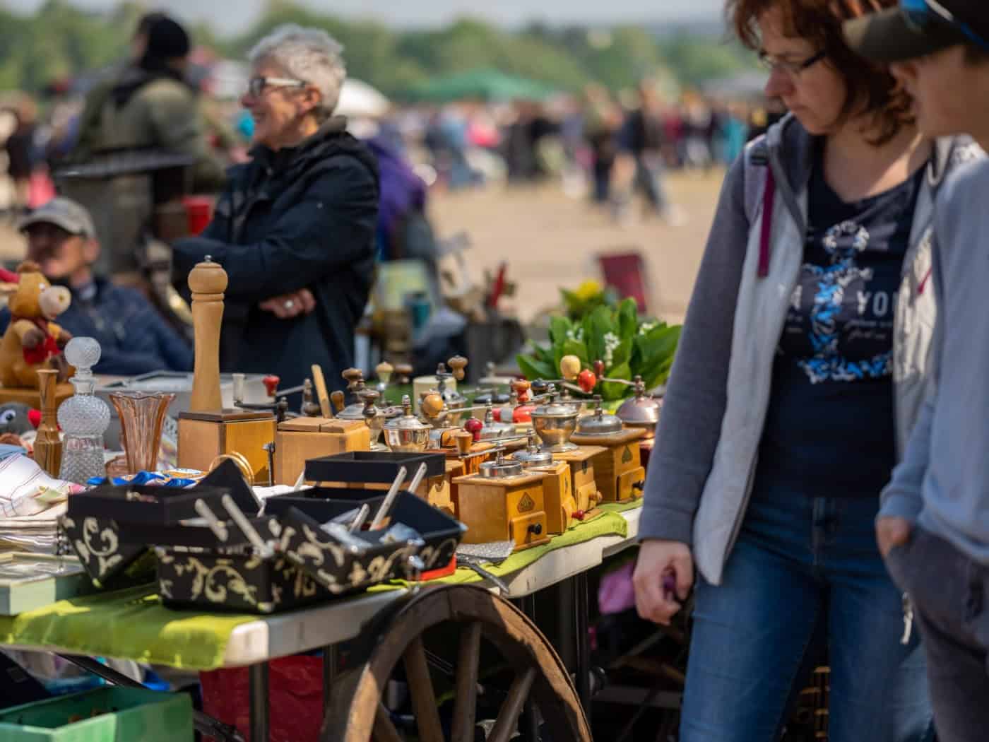 Ausschnitt eines Flohmarktstandes auf dem Elbeflohmarkt in Dresden mit historischen Kaffeemühlen, Holzkästchen und Dekorationen; rechts im Vordergrund sind unscharf zwei Besucher beim Betrachten der Waren zu sehen.