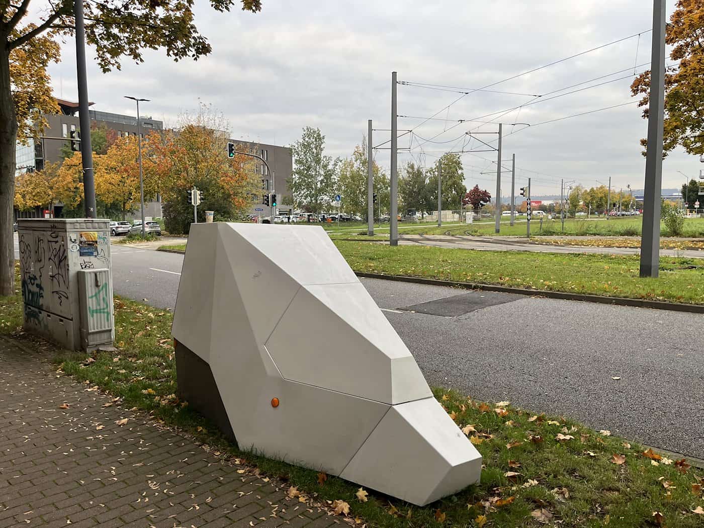 Schrägansicht des futuristisch geformten, hellgrauen Blitzer-Anhängers auf dem gepflasterten Gehweg neben der Straße An der Flutrinne; im Hintergrund Grünanlagen, Oberleitungen und herbstliche Bäume unter trübem Himmel.