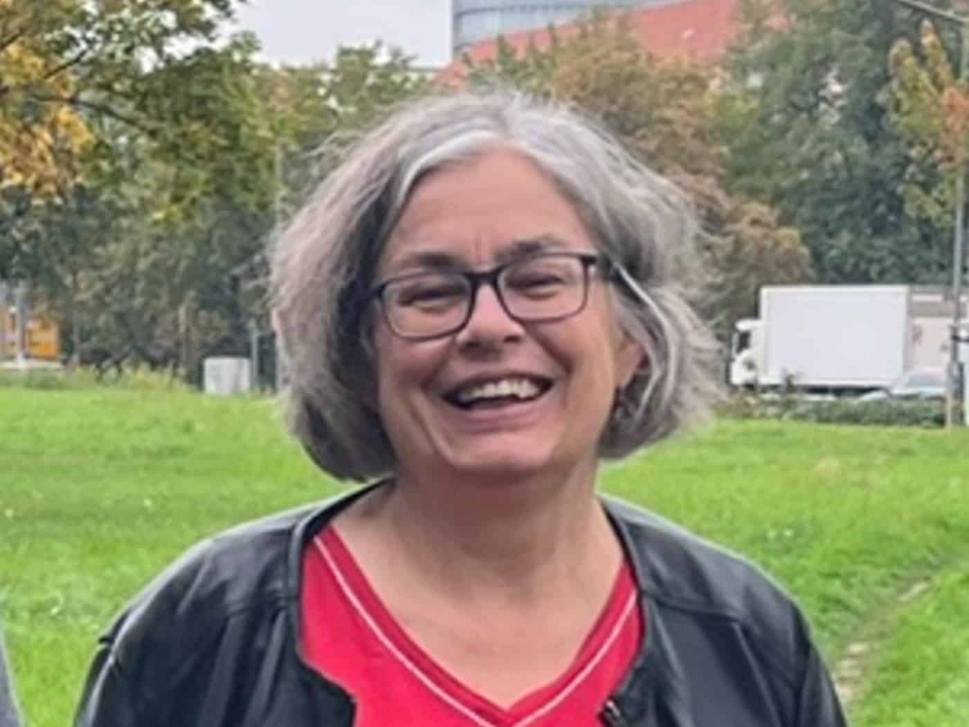 Porträt von Eva Jähnigen, Zweite Bürgermeisterin Dresdens. Sie trägt eine Brille, rotes Shirt und schwarze Jacke und lächelt im Freien vor einer grünen Parkfläche.