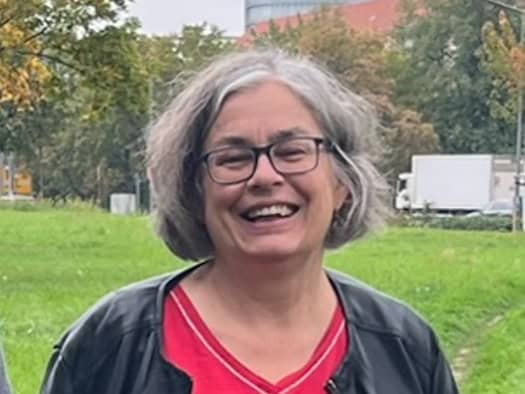 Porträt von Eva Jähnigen, Zweite Bürgermeisterin Dresdens. Sie trägt eine Brille, rotes Shirt und schwarze Jacke und lächelt im Freien vor einer grünen Parkfläche.