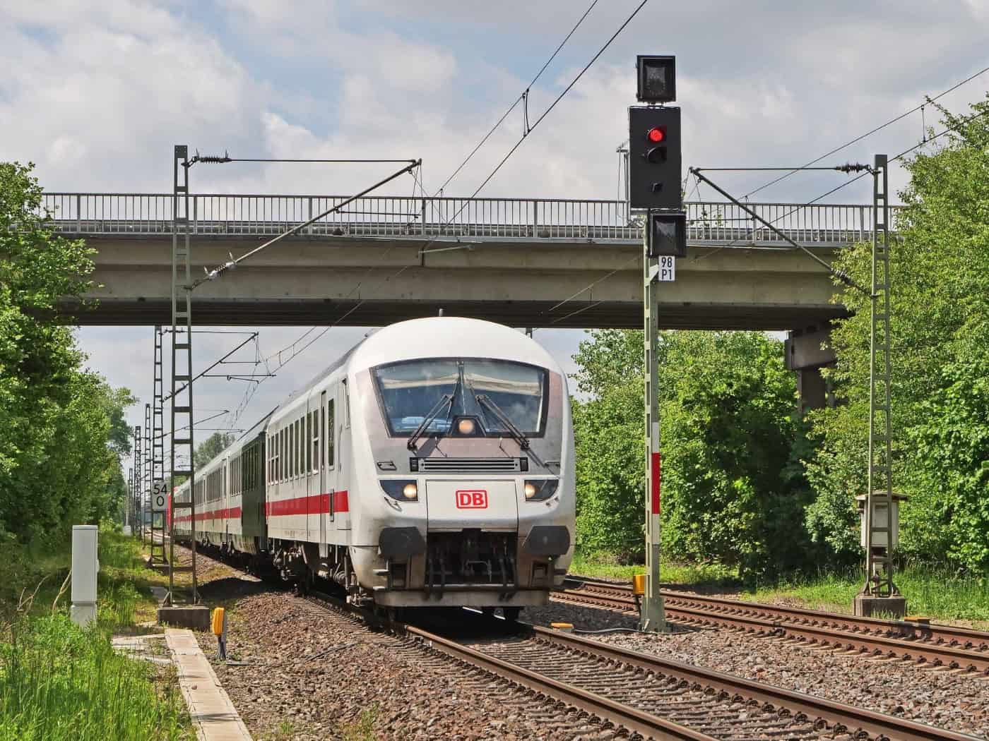 Ein weiß-roter Intercity-Zug der Deutschen Bahn fährt auf mehrgleisiger Strecke unter einer Straßenbrücke und Signalanlagen durch eine grüne Landschaft.
