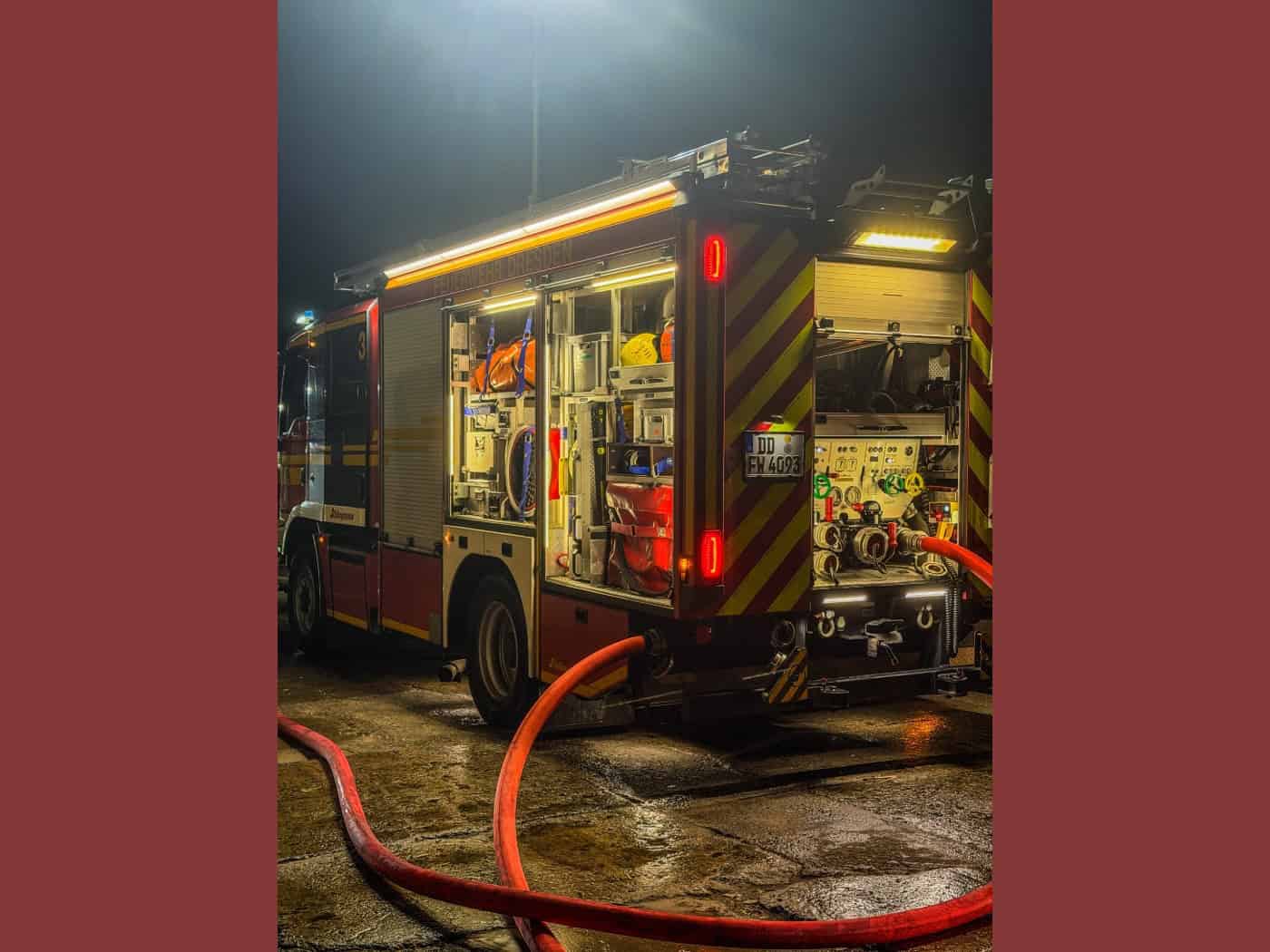 Nächtliche Aufnahme eines beleuchteten Einsatzfahrzeugs der Feuerwehr Dresden mit geöffneten Geräteräumen und Heckklappe. Im Vordergrund verläuft ein roter Löschwasser-Druckschlauch über den nassen, spiegelnden Boden eines Betriebsgeländes.