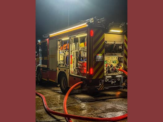 Nächtliche Aufnahme eines beleuchteten Einsatzfahrzeugs der Feuerwehr Dresden mit geöffneten Geräteräumen und Heckklappe. Im Vordergrund verläuft ein roter Löschwasser-Druckschlauch über den nassen, spiegelnden Boden eines Betriebsgeländes.