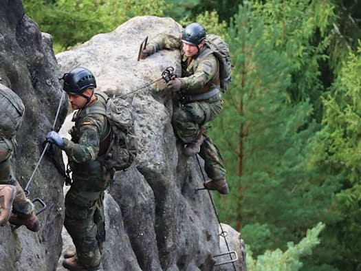 Drei Soldaten in Flecktarn mit Helm und Rucksack klettern gesichert an einem Sandsteinfelsen, im Hintergrund Nadelwald.