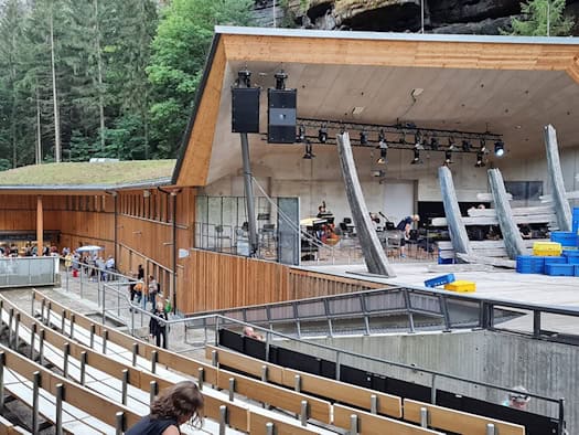 Holzverkleidetes Funktionsgebäude mit überdachtem Orchesterpavillon neben der Bühne der Felsenbühne Rathen, davor Zuschauerbänke, dahinter Wald und Sandsteinfelsen