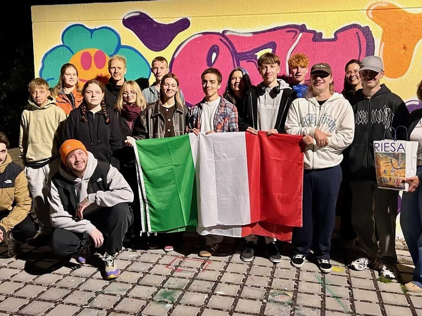Gruppe junger Menschen vor einer Graffiti-Wand hält eine italienische Flagge, eine Frau trägt eine Tüte mit der Aufschrift Riesa