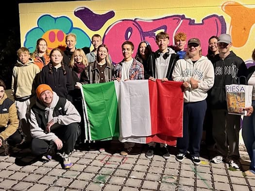Gruppe junger Menschen vor einer Graffiti-Wand hält eine italienische Flagge, eine Frau trägt eine Tüte mit der Aufschrift Riesa