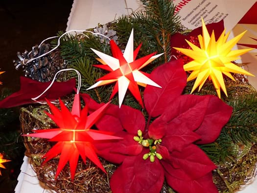 Rote, weiße und gelbe Herrnhuter Sterne leuchten auf einem Adventsgesteck mit Tannengrün und Weihnachtsstern
