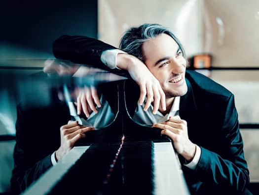 Lächelnder Pianist mit dunklem Anzug stützt sich auf einen Flügel, sein Spiegelbild im Lack