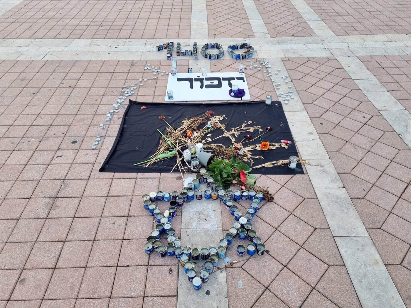 Symbolbild: Ein aus vielen Gedenkkerzen geformter Davidstern auf einem gepflasterten Platz. Dahinter liegen Blumen, ein Schild mit hebräischer Schrift und weitere Anordnungen aus Kerzen.