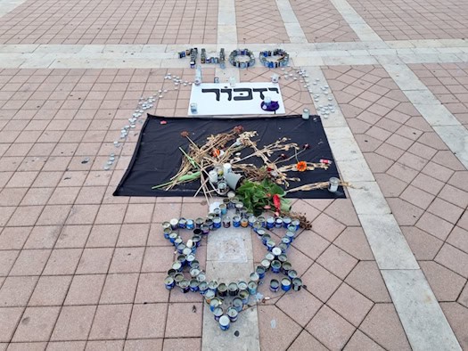Symbolbild: Ein aus vielen Gedenkkerzen geformter Davidstern auf einem gepflasterten Platz. Dahinter liegen Blumen, ein Schild mit hebräischer Schrift und weitere Anordnungen aus Kerzen.