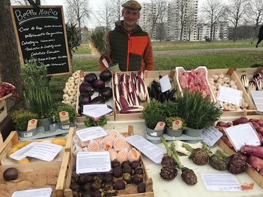 Ein lächelnder Verkäufer mit Mütze steht hinter einem reich gefüllten Marktstand mit Holzkisten voller frischem italienischen Gemüse wie Radicchio, Artischocken, Knoblauch und Kräutertöpfen. Eine Tafel trägt die Aufschrift „Bella Italia“.