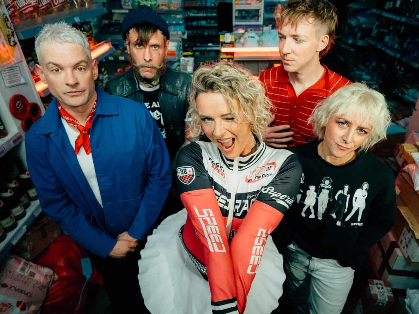Die fünf Bandmitglieder von Jennifer Rostock posieren gemeinsam in einem Kiosk; im Zentrum steht Frontfrau Jennifer Weist mit blonden Haaren und einem auffälligen weiß-roten Outfit, umringt von ihren Bandkollegen.