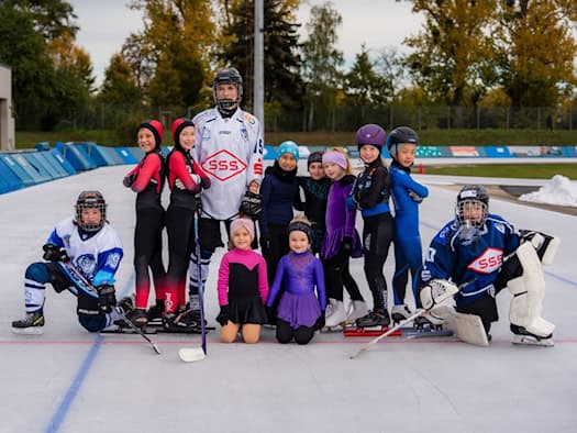 Kinder und Jugendliche verschiedener Eissportdisziplinen in Sportbekleidung, Eishockey-Ausrüstung und Schlittschuhen posieren gemeinsam als Gruppe auf der eisbedeckten Freiluftbahn der JOYNEXT Arena Dresden. Im Hintergrund stehen Bäume und Masten.