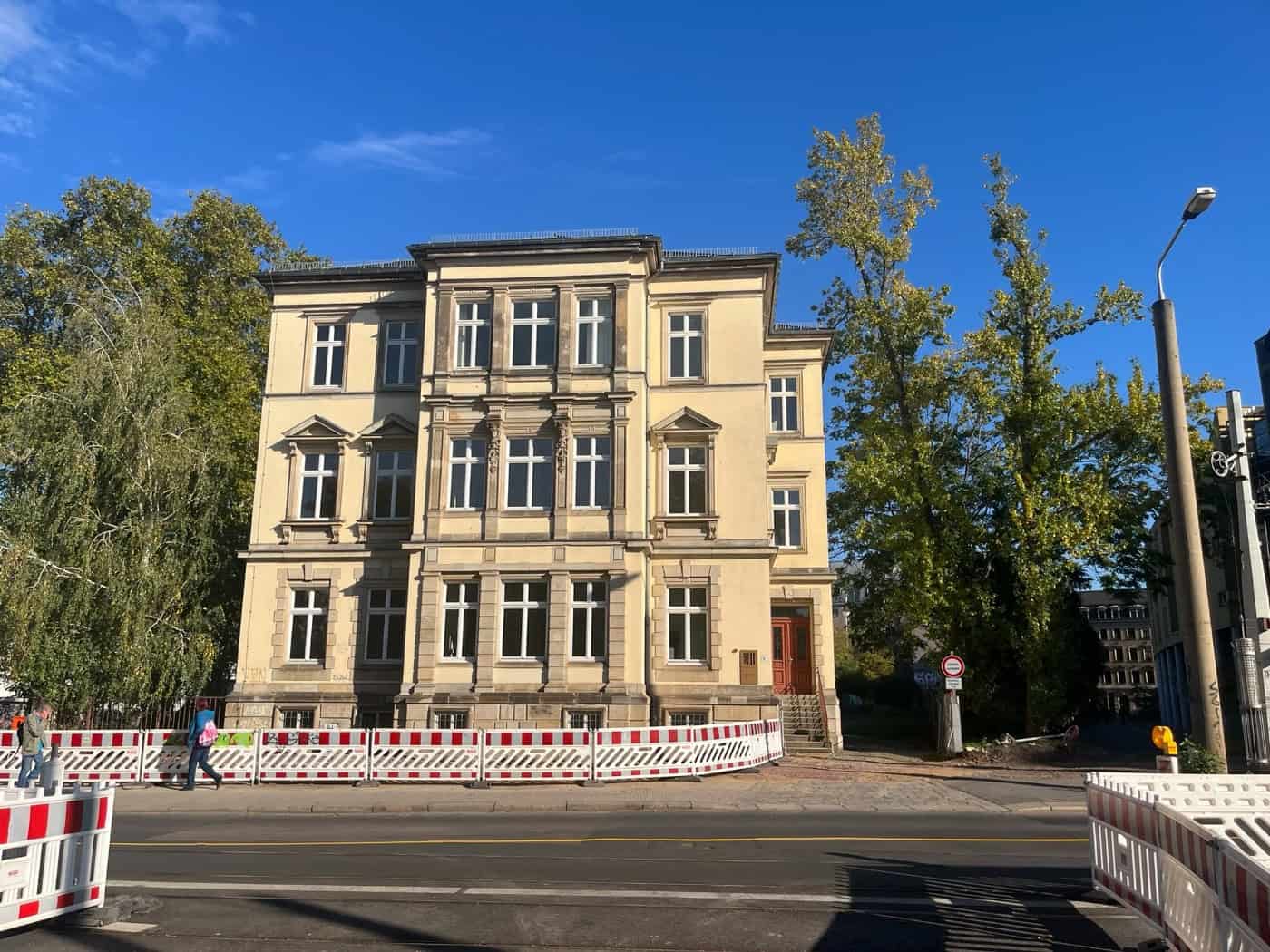 Vorderansicht des sanierten, mehrstöckigen, gelben Gründerzeitgebäudes der Jugendhilfe im Strafverfahren an der Königsbrücker Straße 8 in Dresden; vor dem Haus stehen rot-weiße Bauzäune, daneben wachsen grüne Bäume unter blauem Himmel.