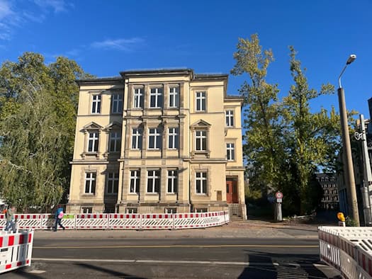 Vorderansicht des sanierten, mehrstöckigen, gelben Gründerzeitgebäudes der Jugendhilfe im Strafverfahren an der Königsbrücker Straße 8 in Dresden; vor dem Haus stehen rot-weiße Bauzäune, daneben wachsen grüne Bäume unter blauem Himmel.